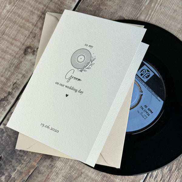 A stack of cream-colored wedding invitation cards with a vinyl record graphic and text, placed on top of a black vinyl record and a beige envelope over a rustic wooden surface.