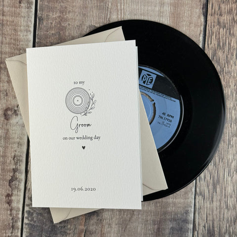 A wedding invitation card with a vinyl record illustration and text, layered over cream envelopes and a black record, displayed on a rustic wooden background.