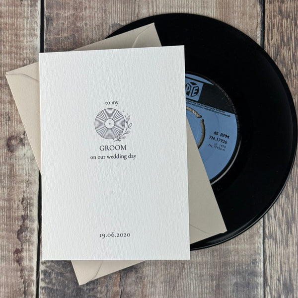 A black vinyl record partially covered by a white printed card and beige envelopes, arranged on a rustic wood plank surface.